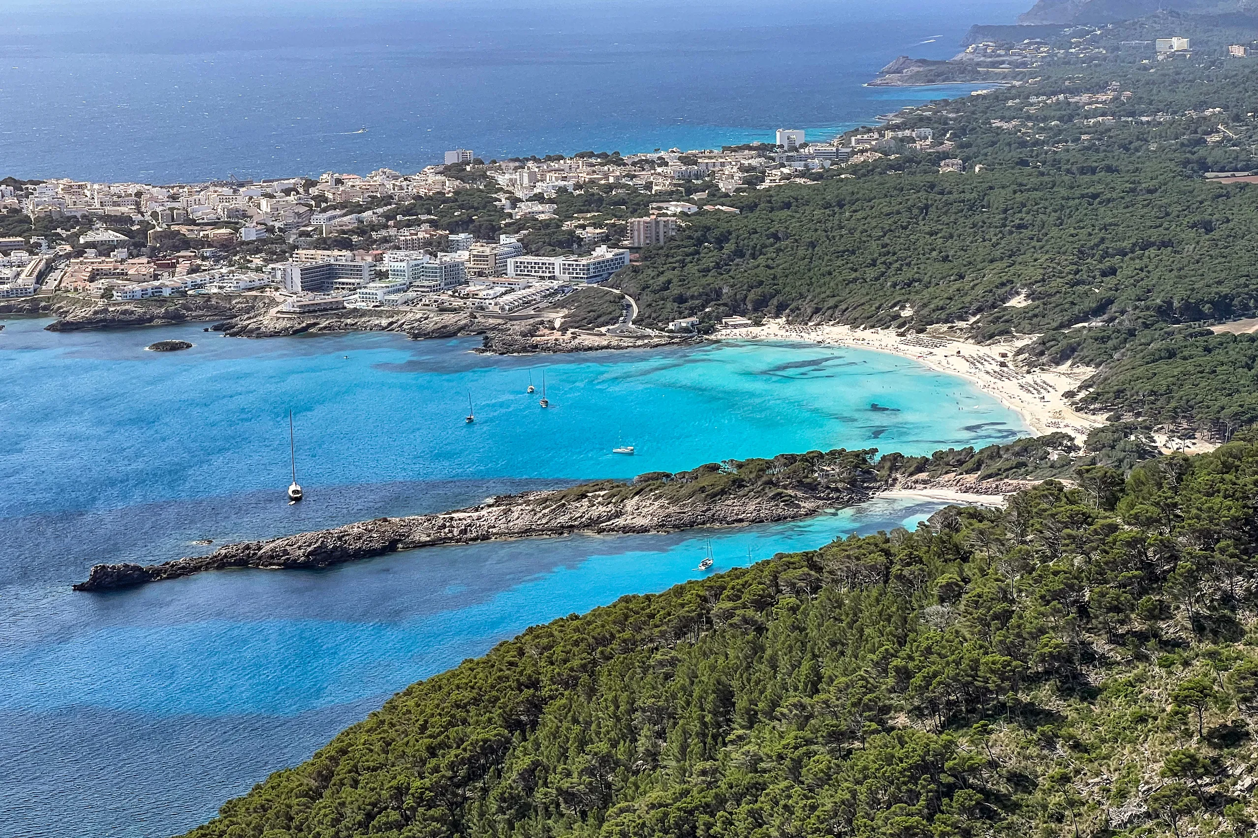Blick von einer bewaldeten Anhöhe auf die Cala Agulla: tiefblaues und türkisfarbenes Wasser, ein heller Sandbogen und dichter Kiefernwald dahinter, im Hintergrund die Häuser von Cala Ratjada.
