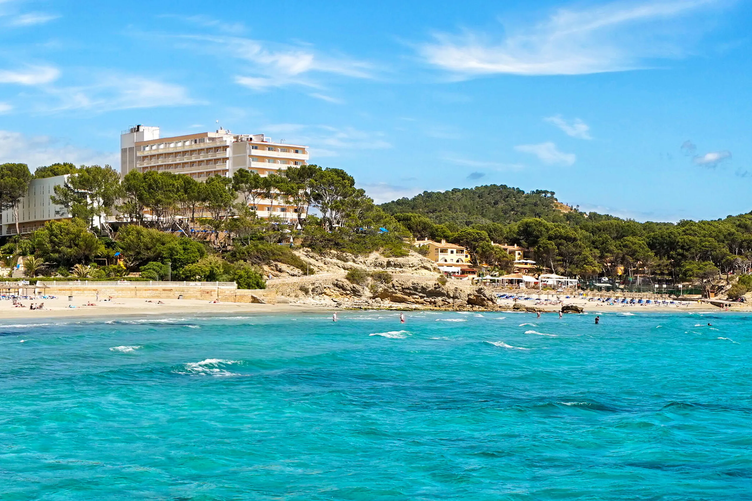 Blick über türkisfarbenes Wasser auf den Sandstrand von Peguera mit Hotelbauten und bewaldeten Felsnasen zu beiden Seiten der Bucht.
