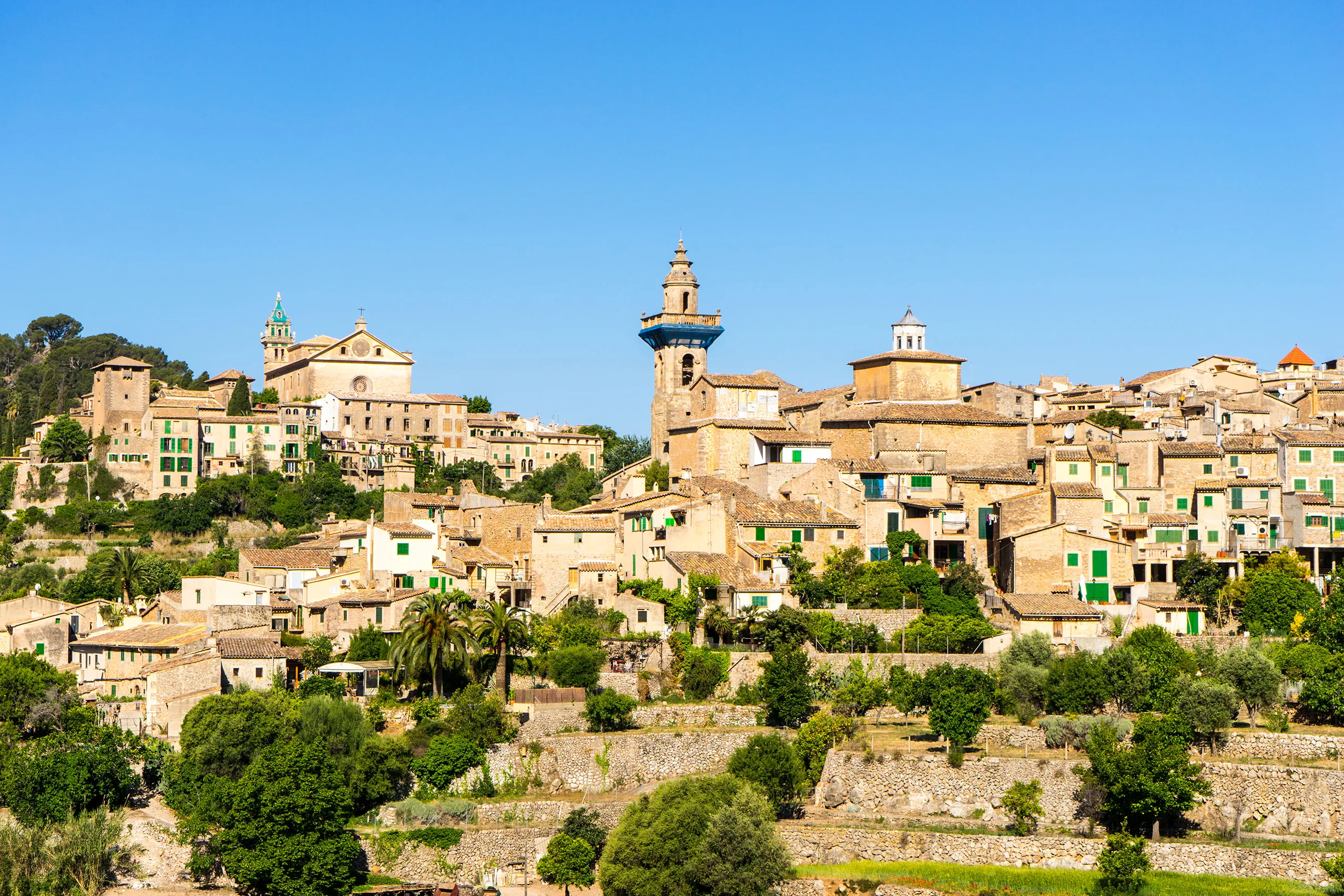 Valldemossa in der Gesamtansicht: dicht gestaffelte Steinhäuser am Hang, darüber die Türme der Kartause und der Kirche, ringsum Terrassen und Bäume.
