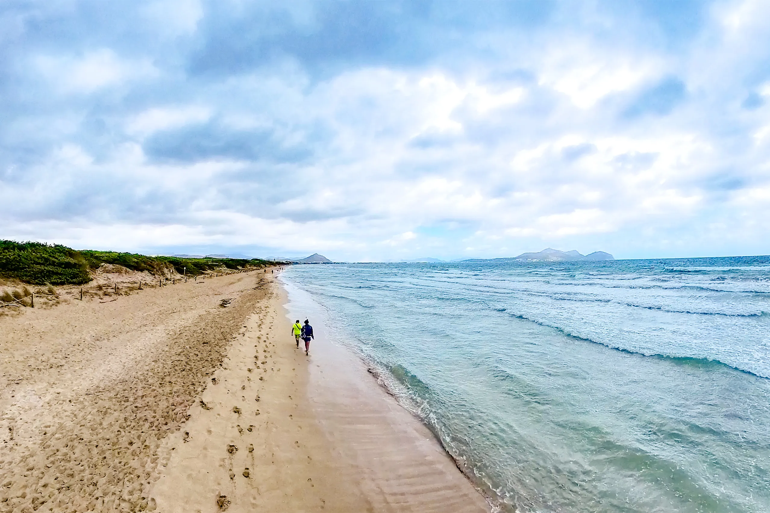 Weiter, fast menschenleerer Sandstrand unter Wolkenhimmel, links eine niedrige bewachsene Düne statt Bebauung, rechts die auflaufende Brandung; zwei Spaziergänger am Wasser, am fernen Horizont die Landzunge von Alcanada und die Berge.
