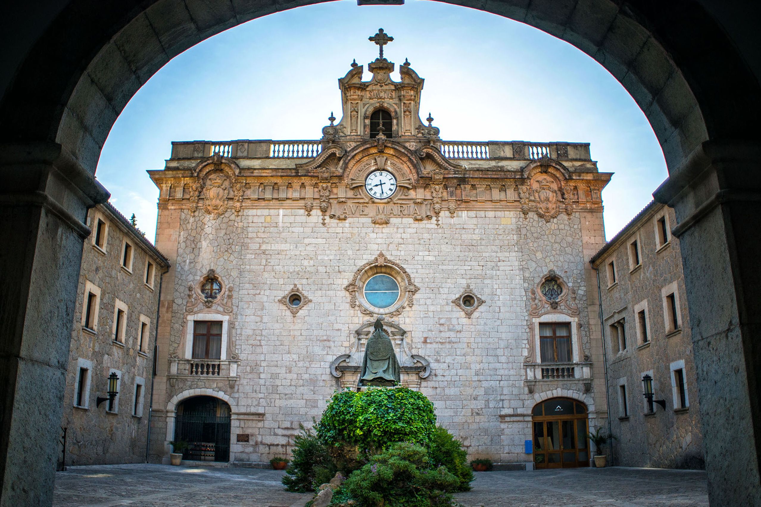 Die barocke Fassade der Klosterkirche von Lluc mit Uhr und Glockengiebel, gesehen durch einen dunklen steinernen Torbogen.
