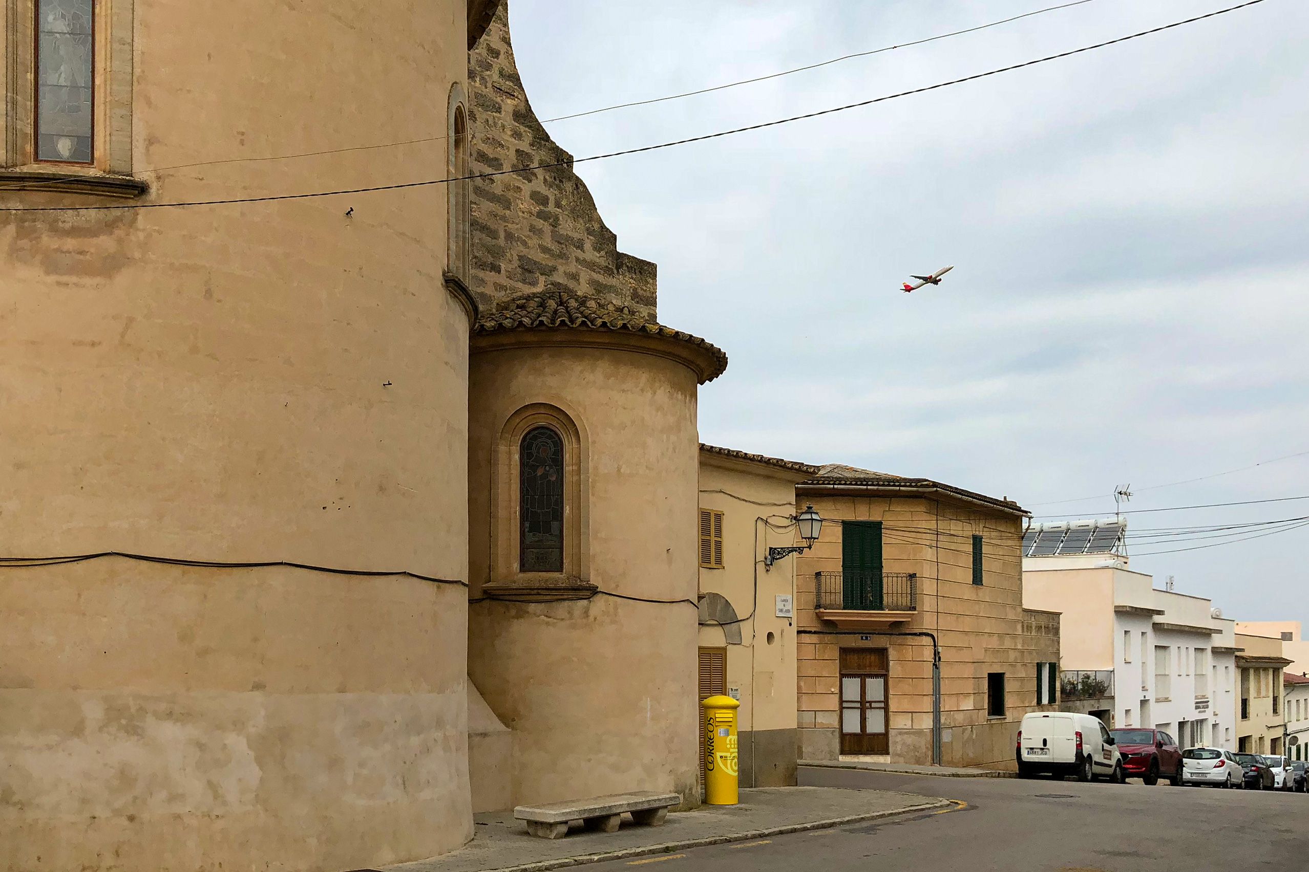 Die Apsis der Kirche von Sant Jordi im Vordergrund, dahinter Dorfhäuser, ein gelber Briefkasten und ein startendes Flugzeug am Himmel.
