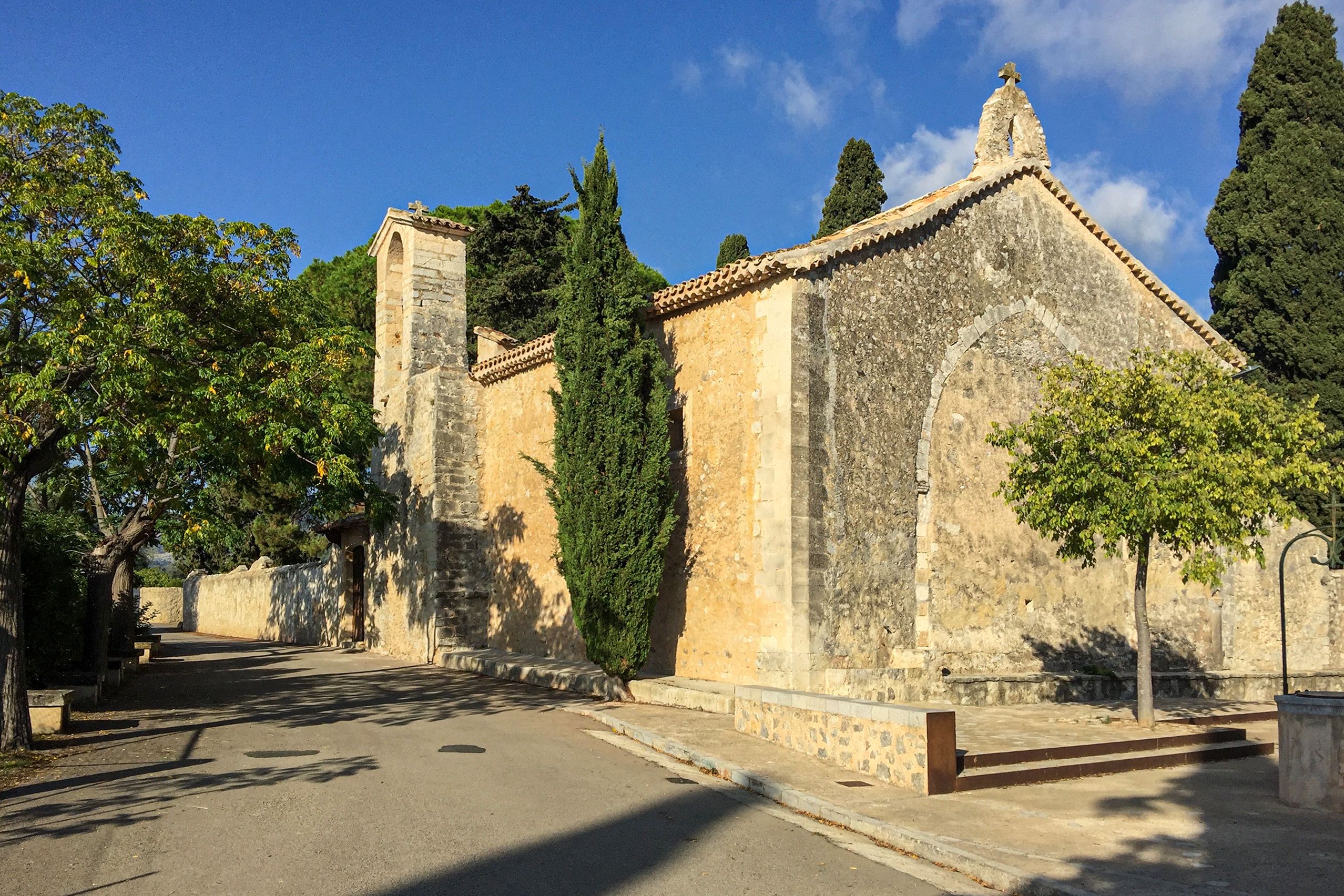 Die alte Kirche Sant Miquel bei Campanet aus hellem Naturstein mit Glockengiebel, flankiert von Zypressen, unter blauem Himmel.
