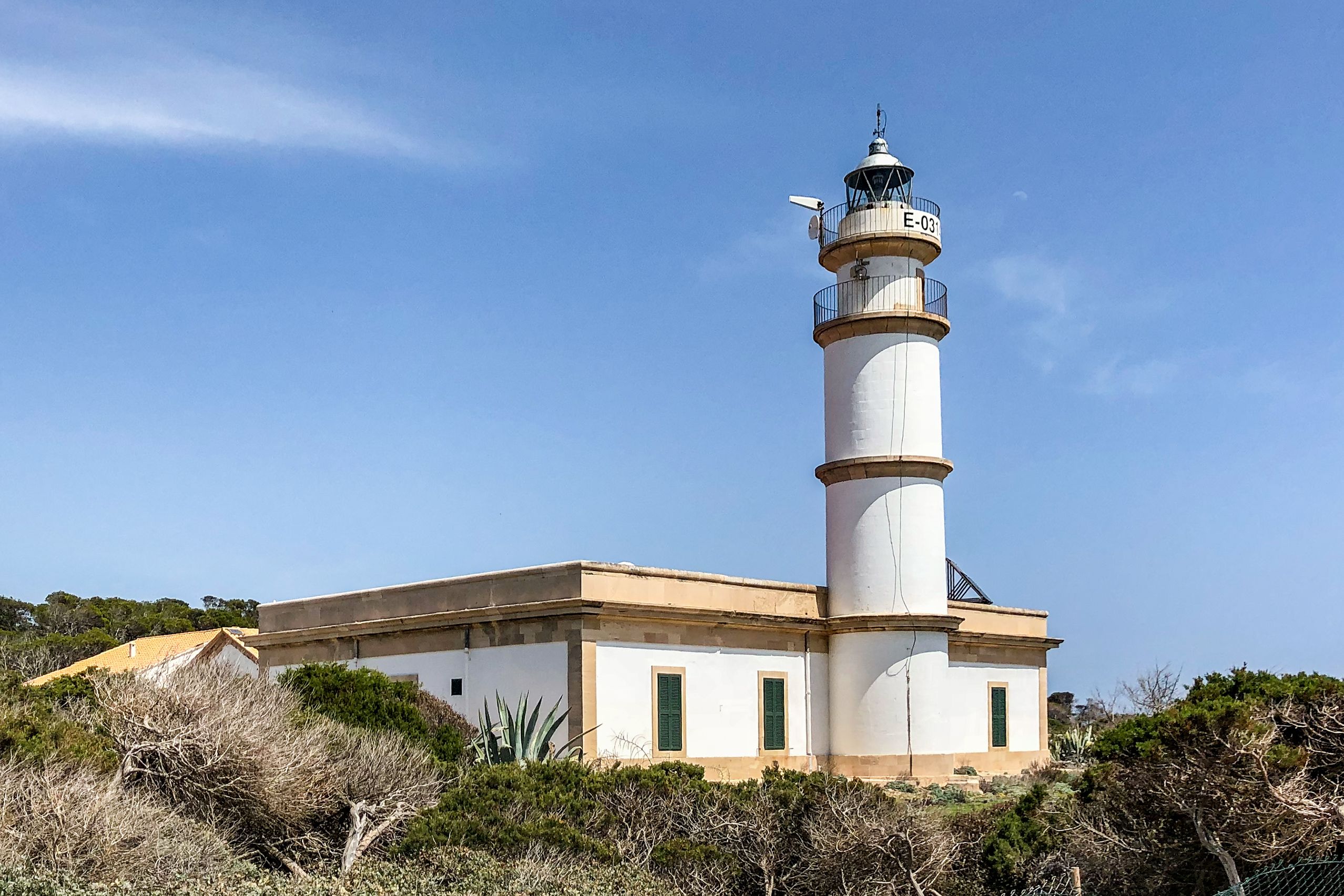 Der weiße Leuchtturm am Cap de ses Salines mit dem niedrigen Wärterhaus, davor zerklüftete helle Felsplatten und das Meer.
