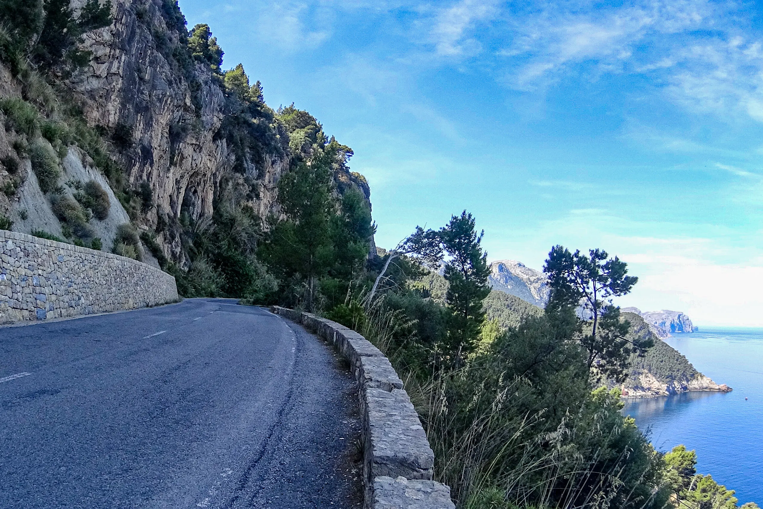 Die Küstenstraße Ma-10 in der Serra de Tramuntana führt in einer Linkskurve am Fels entlang, rechts eine niedrige Trockenmauer, dahinter fällt der Hang zum Meer ab.