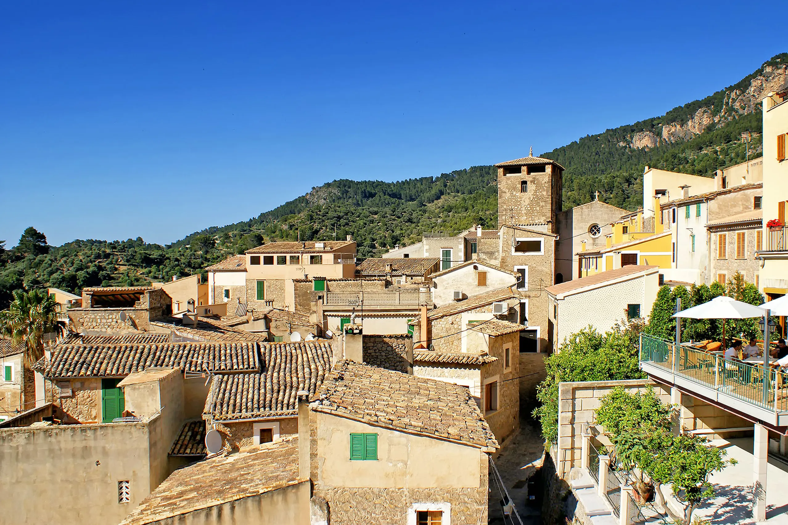 Blick über die Ziegeldächer von Estellencs am Steilhang, dazwischen der wehrhafte Kirchturm, im Hintergrund die bewaldeten Hänge der Tramuntana.
