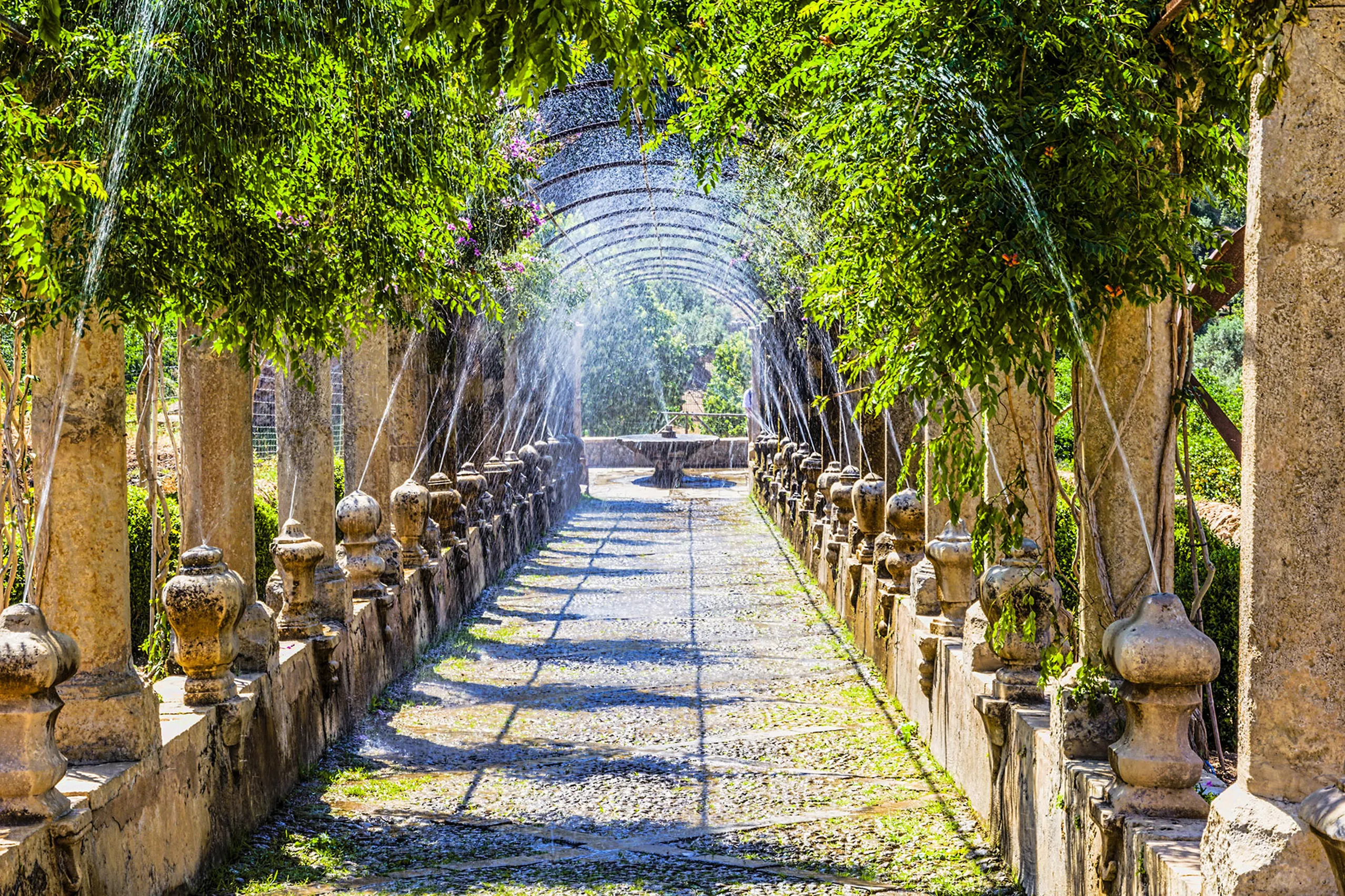 Langer Laubengang in den Jardins d'Alfàbia mit steinernen Säulen und Brüstungen, aus denen feine Wasserstrahlen über den Weg sprühen.
