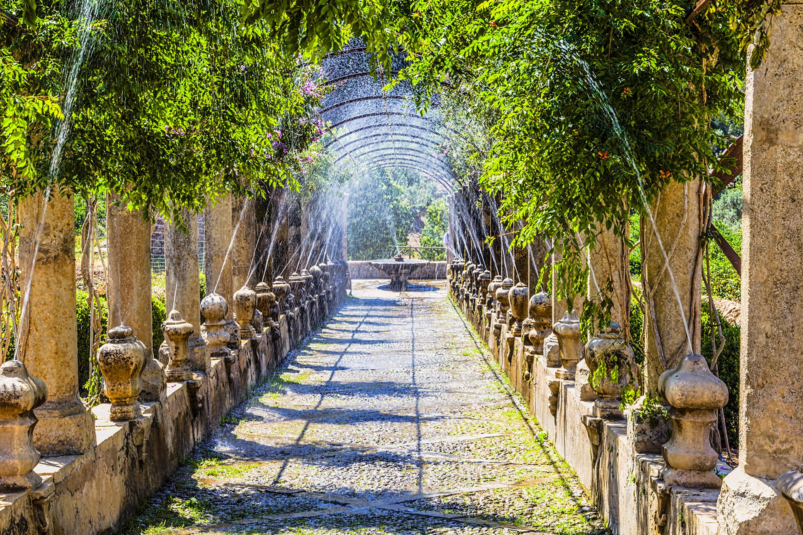 Langer Laubengang in den Jardins d'Alfàbia mit steinernen Säulen und Brüstungen, aus denen feine Wasserstrahlen über den Weg sprühen.
