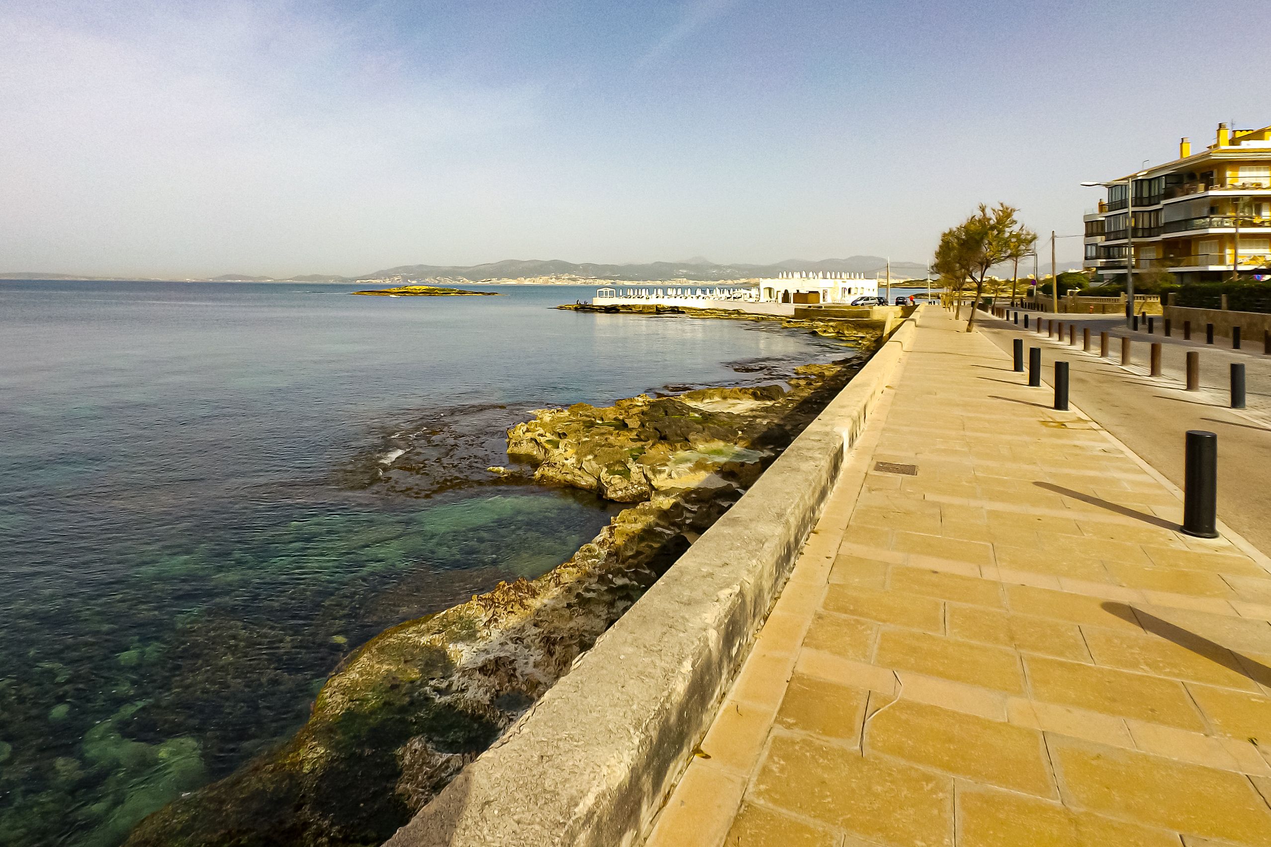 Gepflasterte Uferpromenade in Can Pastilla entlang der flachen Felsküste mit klarem Wasser, dahinter die Bucht von Palma.
