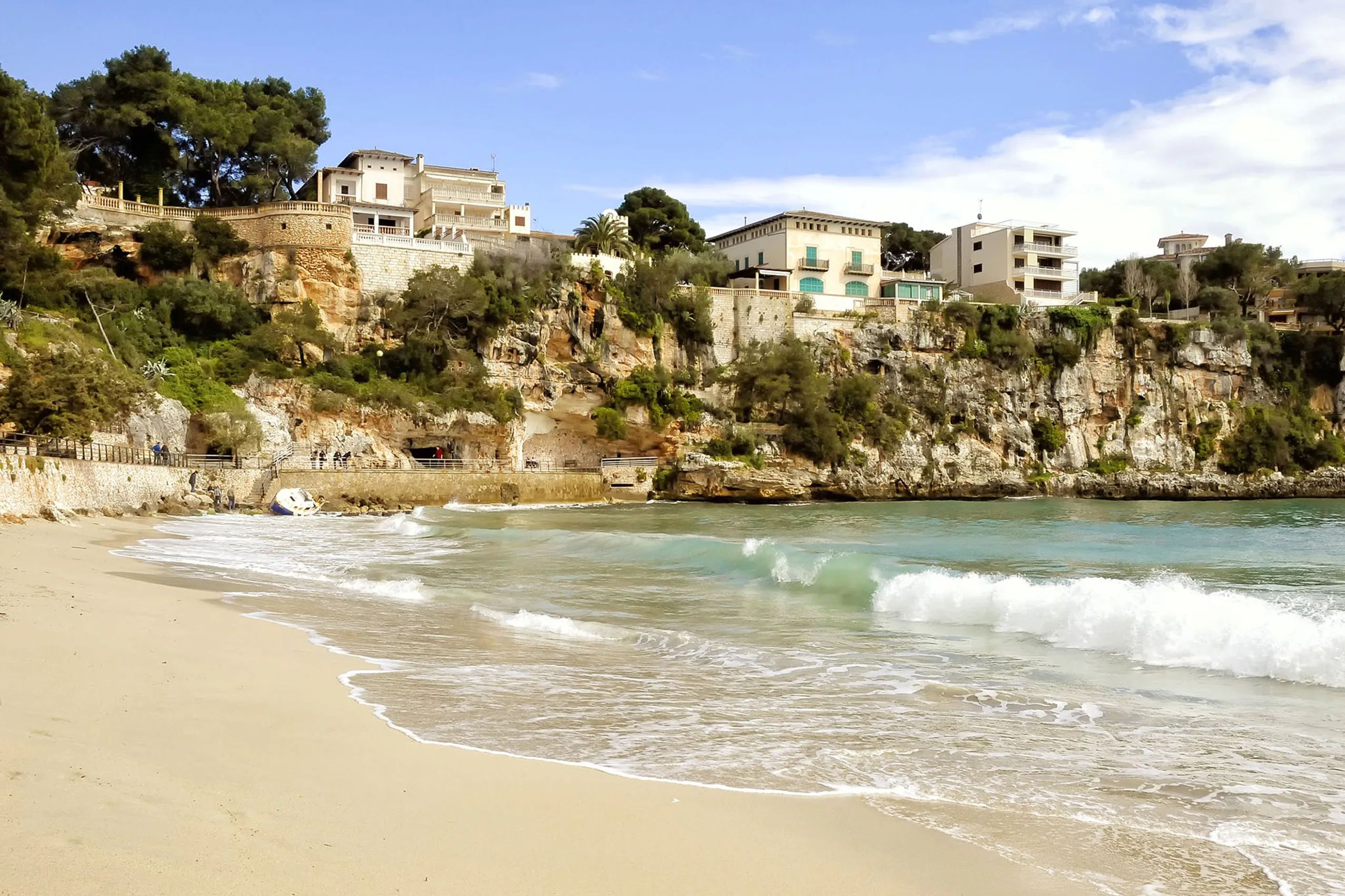 Der Sandstrand von Porto Cristo am Ende der geschützten Bucht mit türkisfarbenem Wasser, Badegästen und der Hotel- und Häuserzeile am Hang dahinter.
