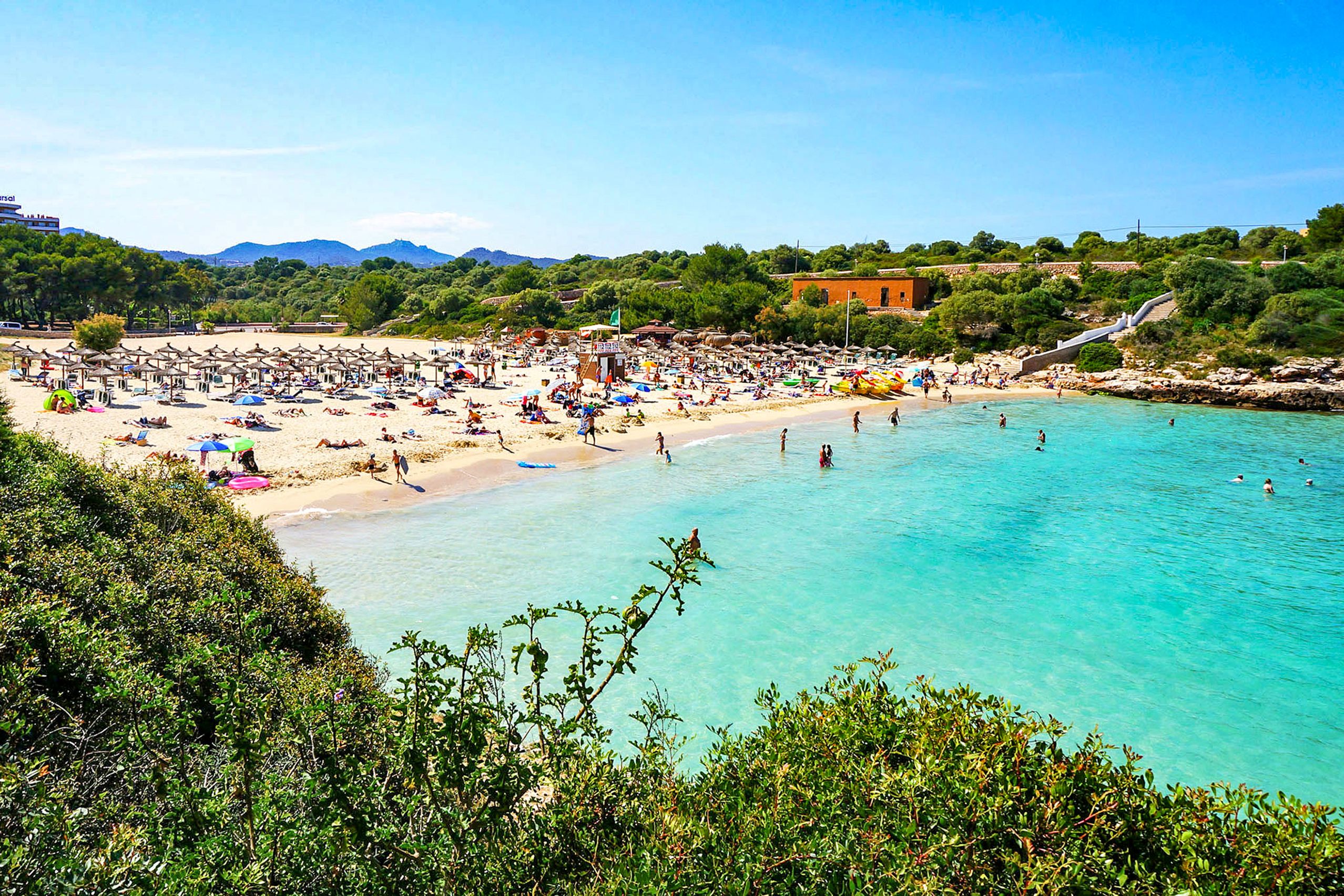 Dicht belegter goldgelber Sandstrand der Cala d'en Marçal mit Schirmreihen und Badegästen, davor sehr klares türkisfarbenes Wasser.
