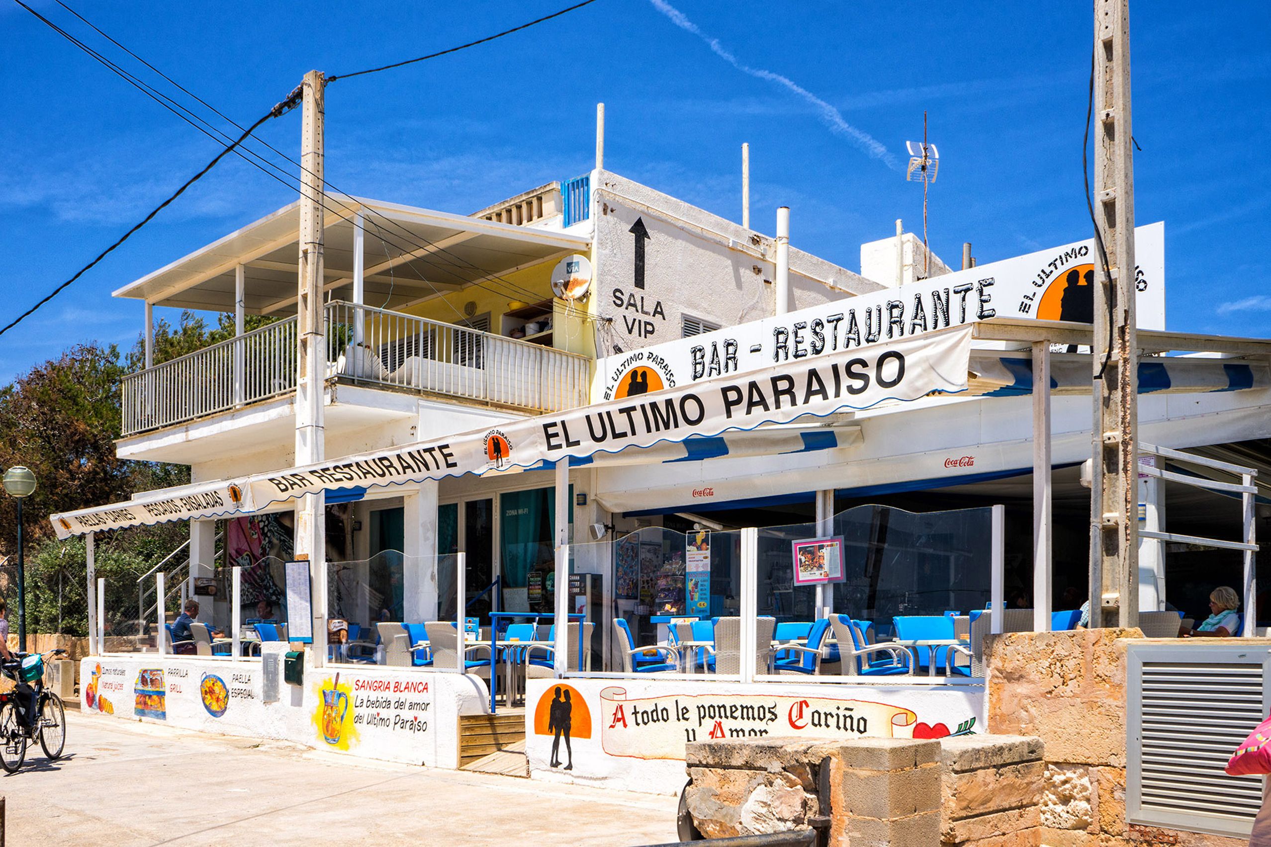 Zweigeschossiges weißes Strandlokal in Ses Covetes mit Terrasse, blauen Stühlen und großen handgemalten Schildern unter blauem Himmel.
