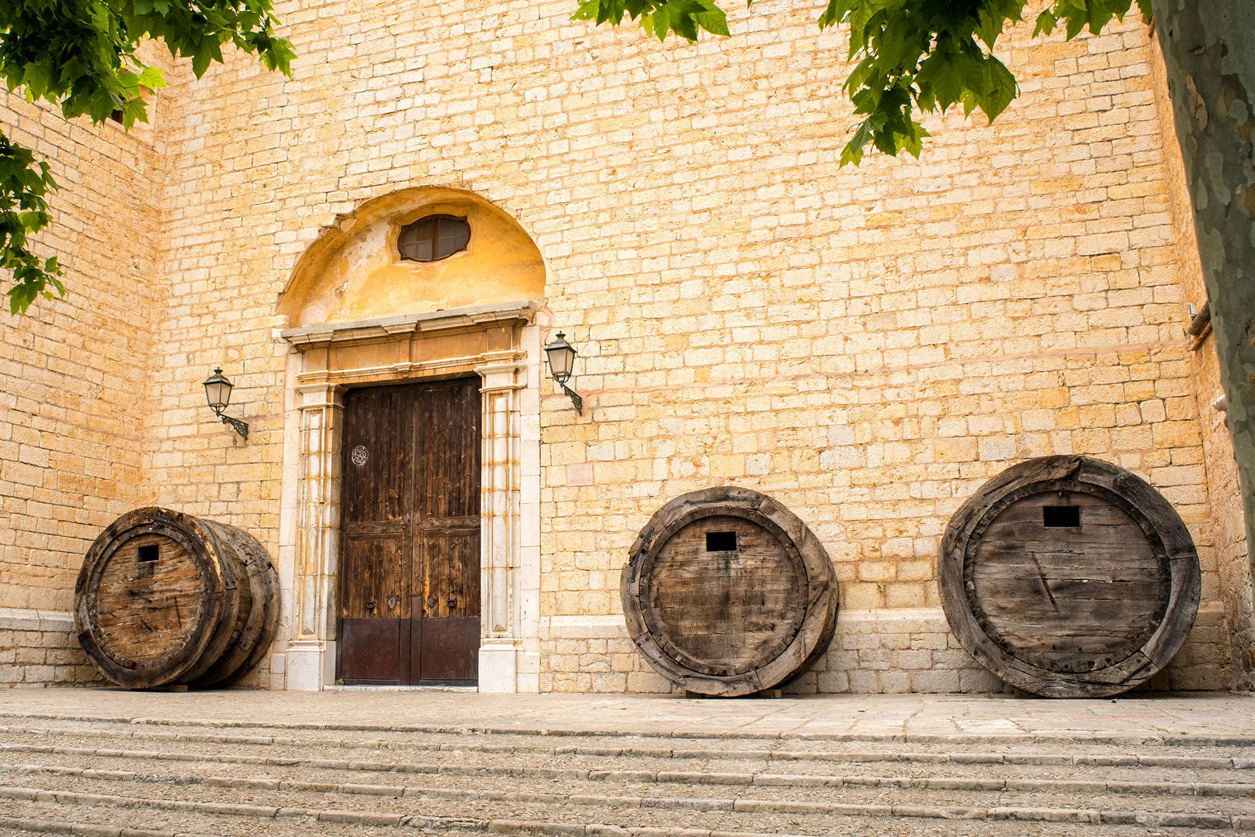 Drei große hölzerne Weinfässer, die an der Sandsteinwand der Kirche von Binissalem neben einem steinernen Portal stehen, davor Stufen.
