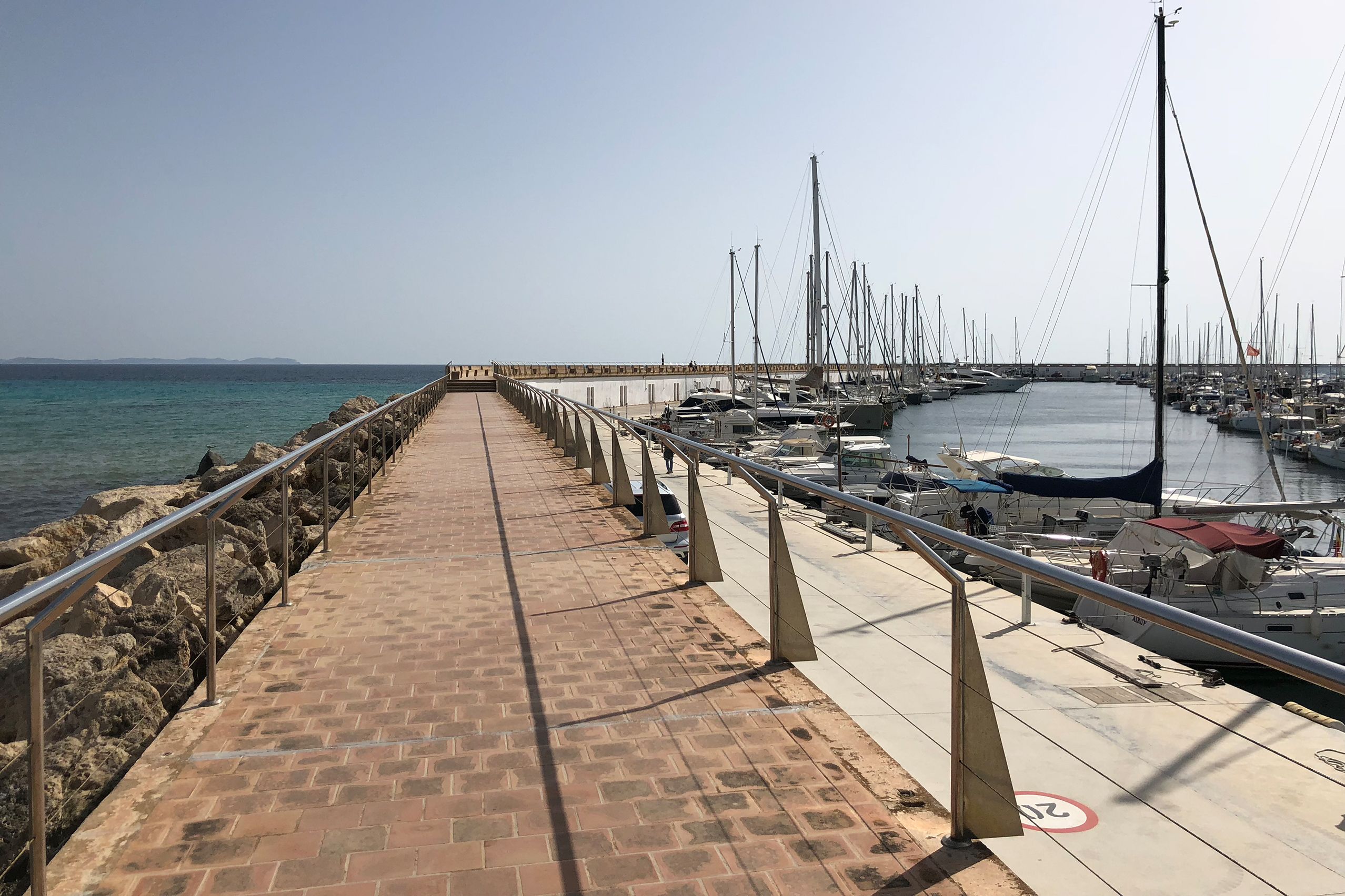 Lange gepflasterte Mole im Hafen von Sa Ràpita mit Geländer, rechts die Stege mit Segelbooten, links die Steinschüttung.
