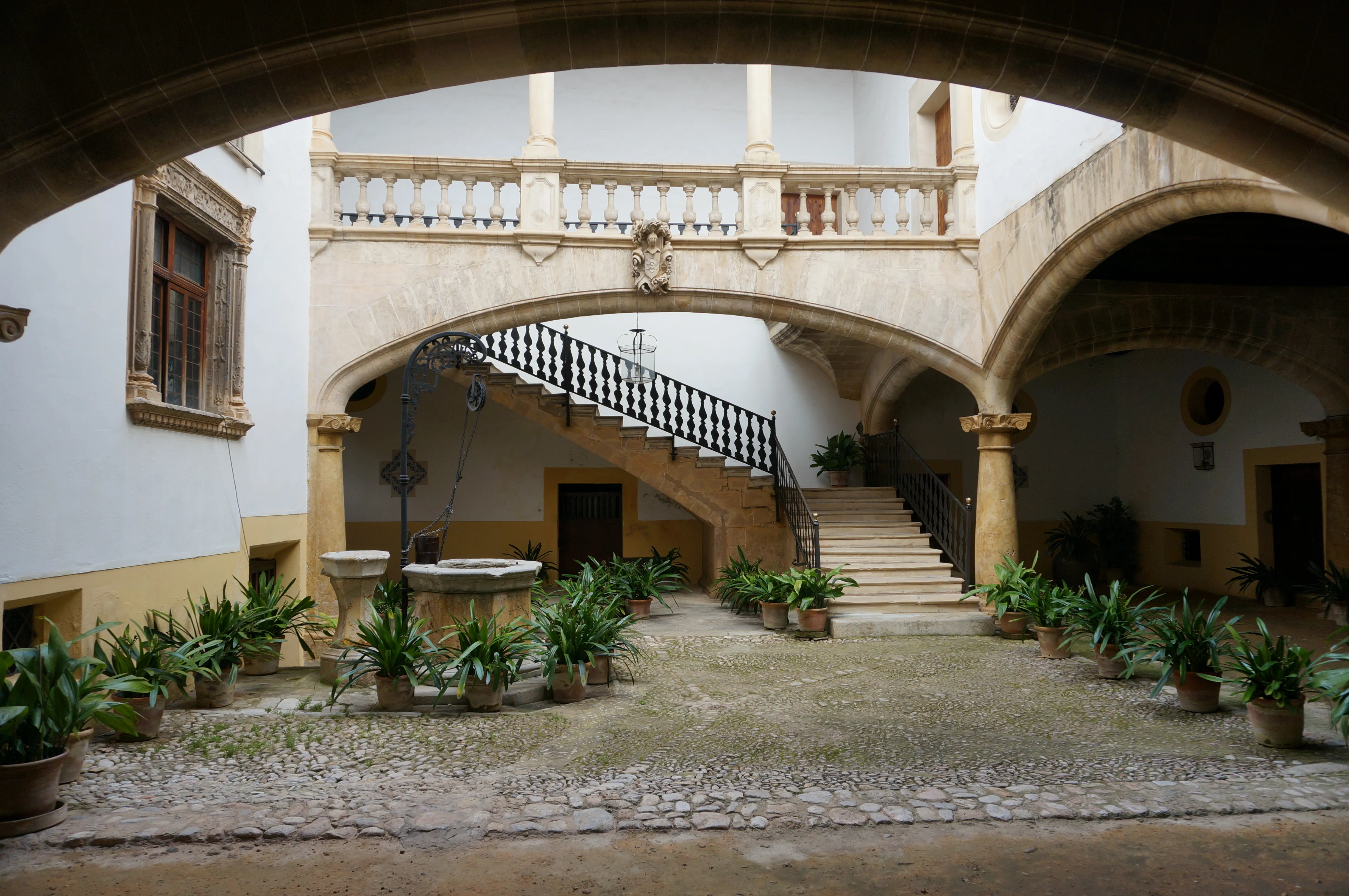 Innenhof eines Stadtpalastes in Palma mit Rundbögen, steinerner Freitreppe, schmiedeeisernem Geländer und Topfpflanzen auf Kopfsteinpflaster.

