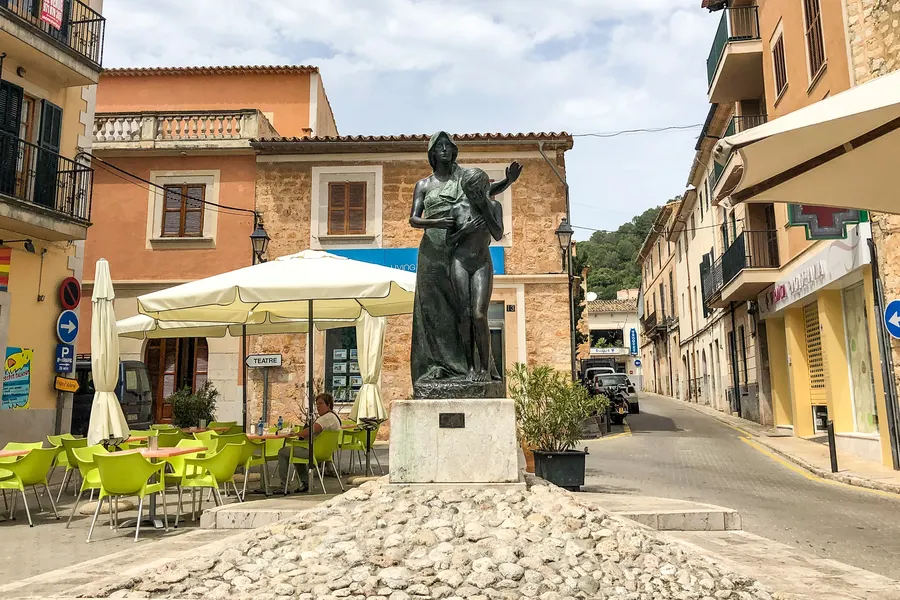Bronzedenkmal einer stehenden Figur auf gepflastertem Sockel an einem Platz in Alaró, ringsum Café-Terrassen mit grünen Stühlen.