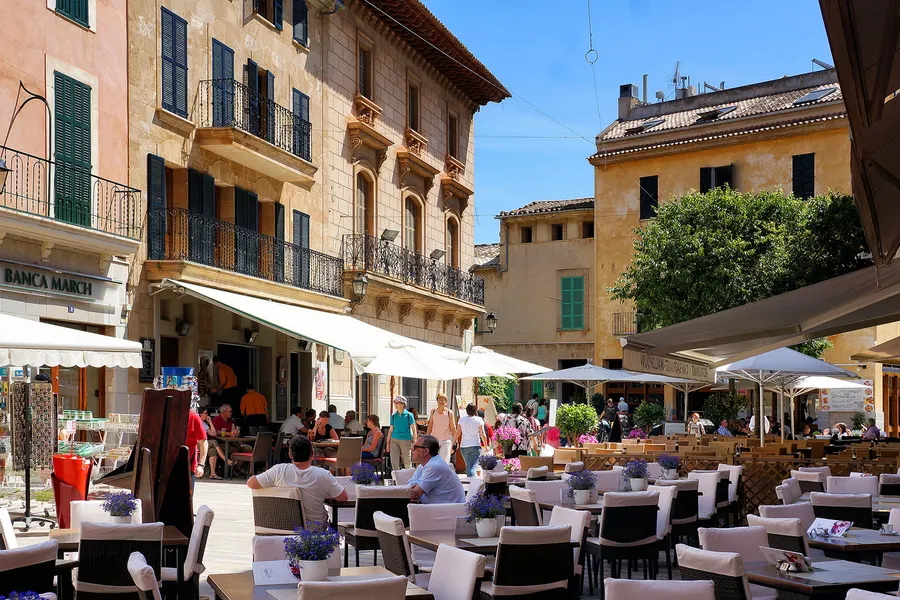 Belebter Platz in Alcúdia mit Straßencafés unter weißen Sonnenschirmen, Gästen an den Tischen und Bürgerhäusern mit schmiedeeisernen Balkonen.
