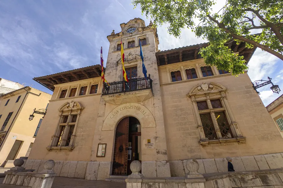 Das Rathaus von Alcúdia, ein Sandsteinbau mit Balkon, Fahnen und Rundbogenportal, davor ein Baum und die gepflasterte Gasse.
