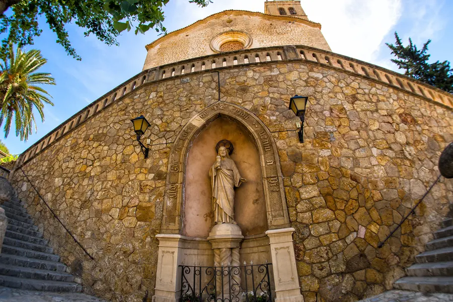Hohe Bruchsteinmauer unterhalb der Kirche von Andratx mit einer Bogennische, in der eine steinerne Heiligenfigur steht, links und rechts Treppen.
