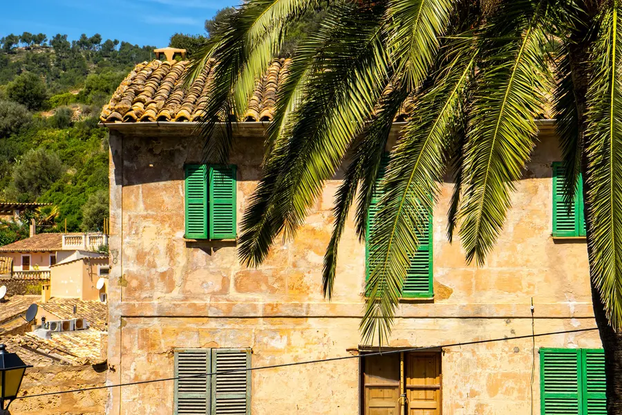 Sandsteinhaus in Andratx mit grünen Fensterläden und Ziegeldach, davor die Wedel einer Palme, im Hintergrund weitere Dächer.
