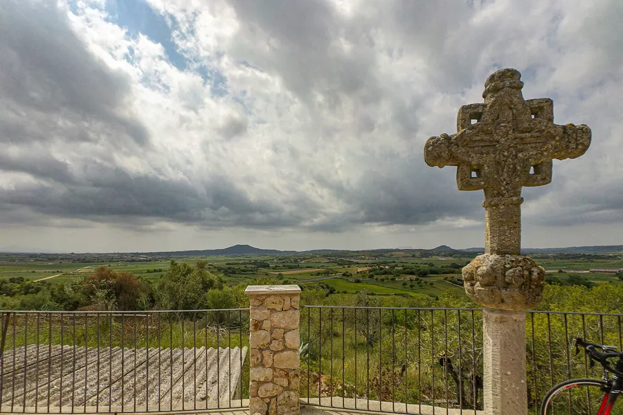 Blick vom Mirador de sa Creu über die weite Ebene des Pla mit Feldern und fernen Hügeln, rechts ein verwittertes Steinkreuz.
