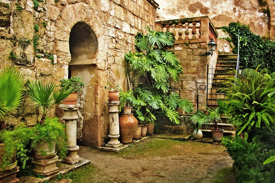 Begrünter Innengarten mit alter Steinmauer, einer Bogennische, Amphoren, Palmen und einer Außentreppe mit Eisengeländer.