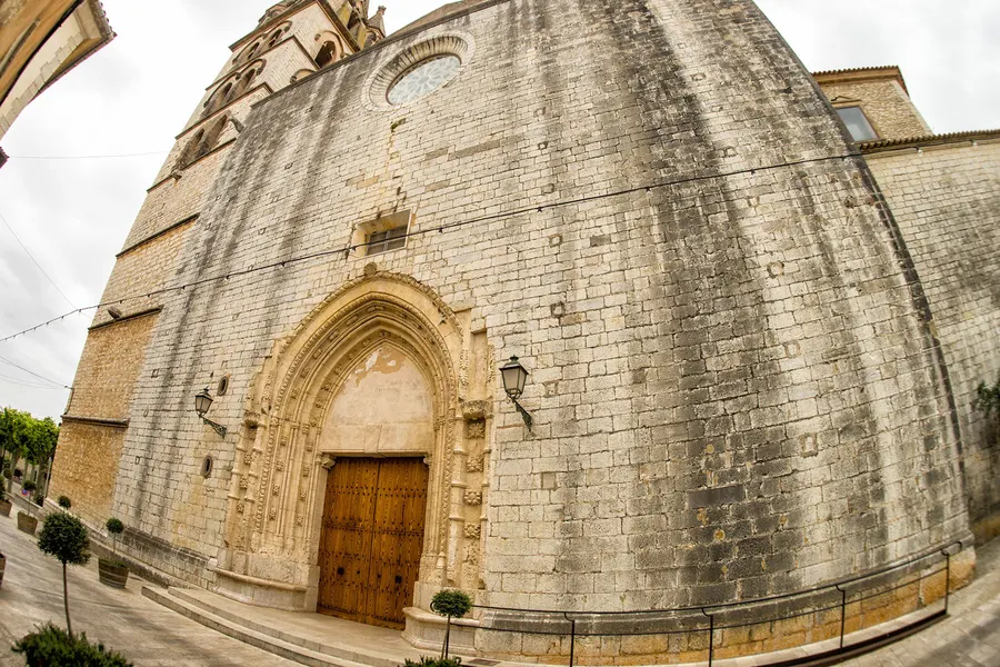 Die wuchtige Kirche von Binissalem aus hellem Sandstein mit Strebepfeilern und gotischem Portal, von schräg unten aufgenommen.
