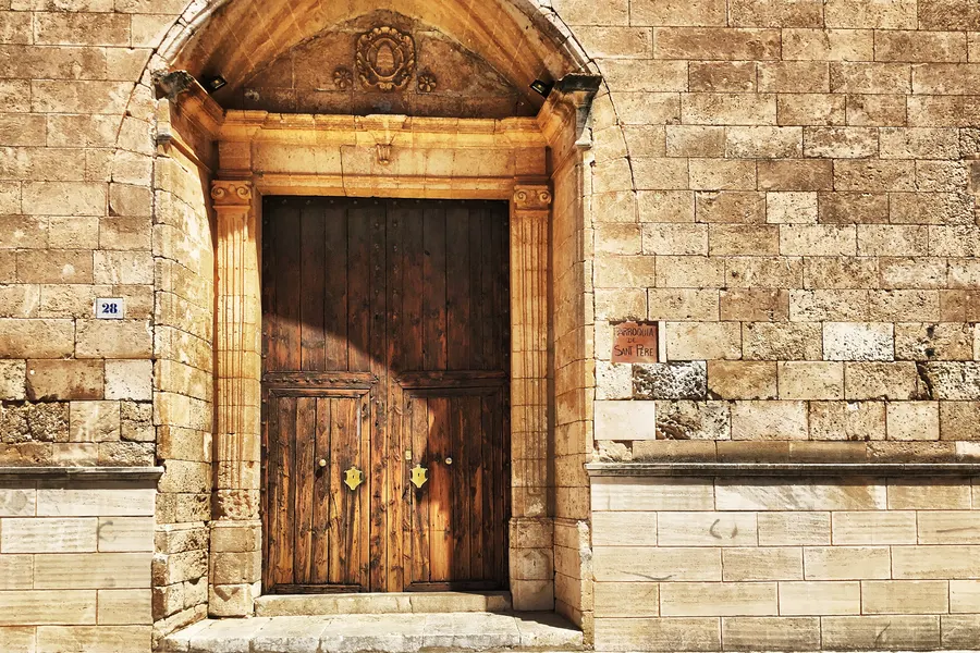 Schweres zweiflügeliges Holzportal der Kirche Sant Pere in Búger, gerahmt von einem schlichten Steingewände aus hellem Sandstein.