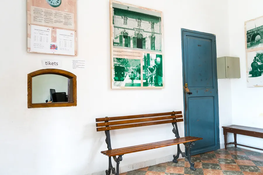 Schalterhalle im Bahnhof Bunyola mit Fahrplänen an der weißen Wand, hölzerner Bank, kleinem Ticketfenster und blauer Tür.
