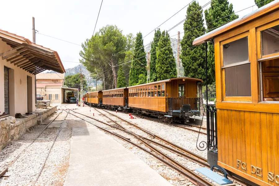 Zug aus hölzernen Wagen am Bahnsteig von Bunyola, daneben das Stationsgebäude mit Vordach, dahinter Zypressen und Kiefern.
