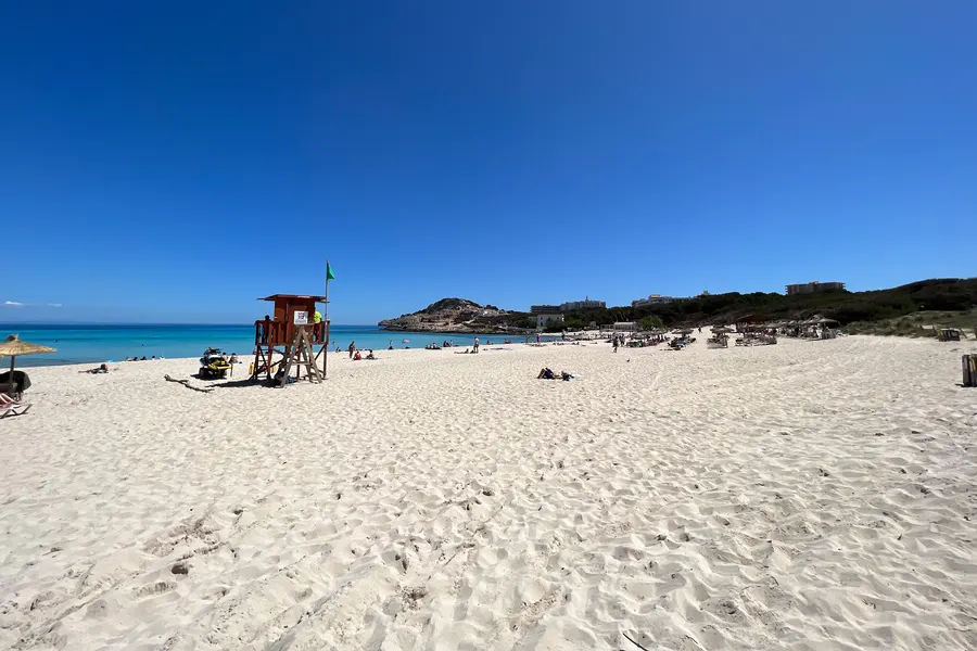 Hölzerner Rettungsschwimmerturm mit grüner Flagge im hellen Sand der Cala Agulla, dahinter Badegäste und das blaue Meer.
