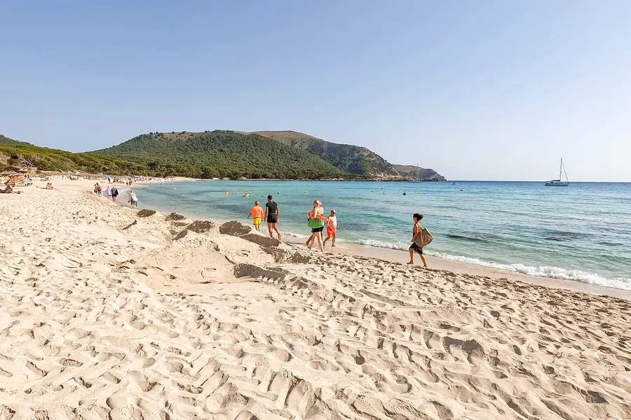 Badegäste auf dem breiten Sandstrand der Cala Agulla, im Hintergrund der bewaldete Hügel und türkisfarbenes Wasser.
