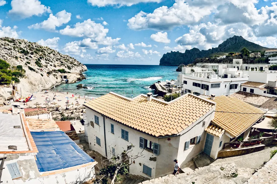 Weiße Häuser mit Ziegeldächern über einer Bucht von Cala de Sant Vicenç, unten Badegäste am Strand, dahinter das offene Meer und eine Landzunge.