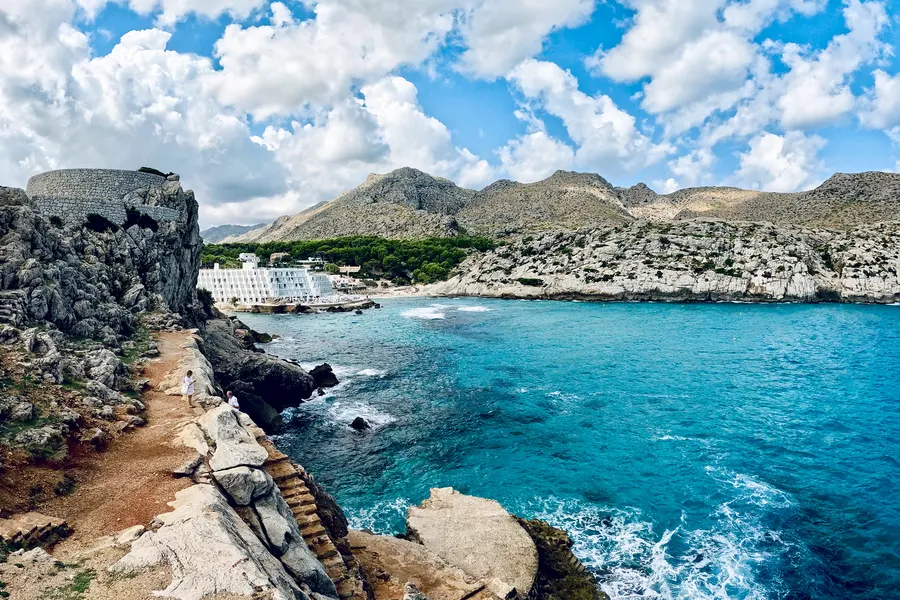 Felsige Uferzone von Cala de Sant Vicenç mit einer in den Stein gehauenen Treppe zum Wasser, kräftig türkisfarbenes Meer und Klippen im Hintergrund.