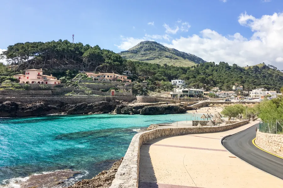 Uferstraße von Cala de Sant Vicenç entlang einer Mauer über dem türkisfarbenen Wasser, dahinter Häuser am bewaldeten Hang und die Berge.