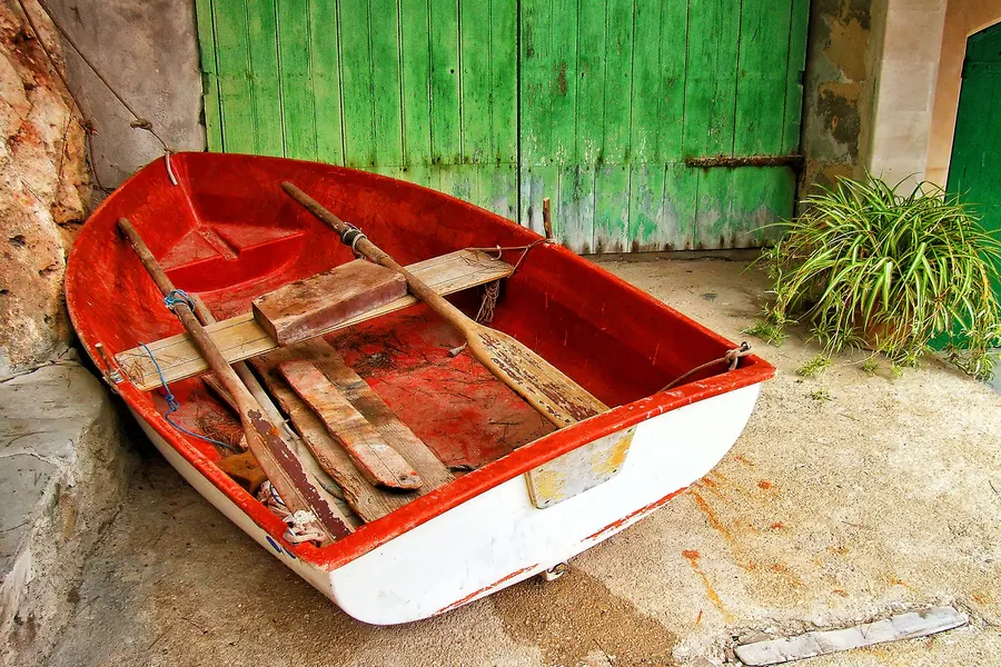 Rot-weißes hölzernes Ruderboot an Land vor einem großen grünen Holztor eines Bootshauses in Cala Figuera.
