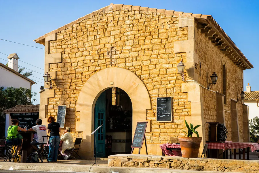 Ehemalige Kirche von Cala Figuera aus Sandstein mit großem Rundbogentor, heute ein Restaurant, davor Tafeln und Gäste.
