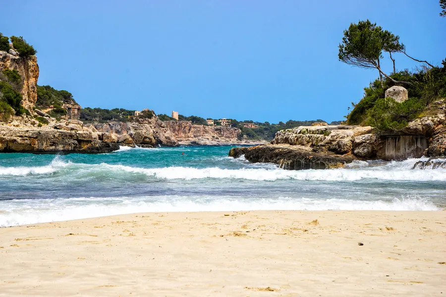Weiße Brandung läuft auf den leeren Sandstrand der Cala Llombards, dahinter die Felswand mit niedrigem Bewuchs.
