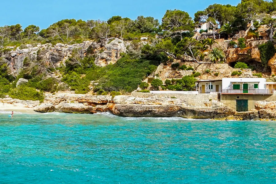 In den Fels gebaute weiße Fischerhäuser mit grünen Holztoren am Rand der Cala Llombards, davor türkisfarbenes Wasser.