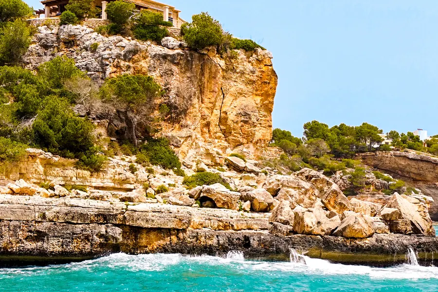 Steile, zerklüftete Sandsteinwand am Rand der Cala Llombards mit Kiefern auf der Kante und aufschäumendem Wasser darunter.
