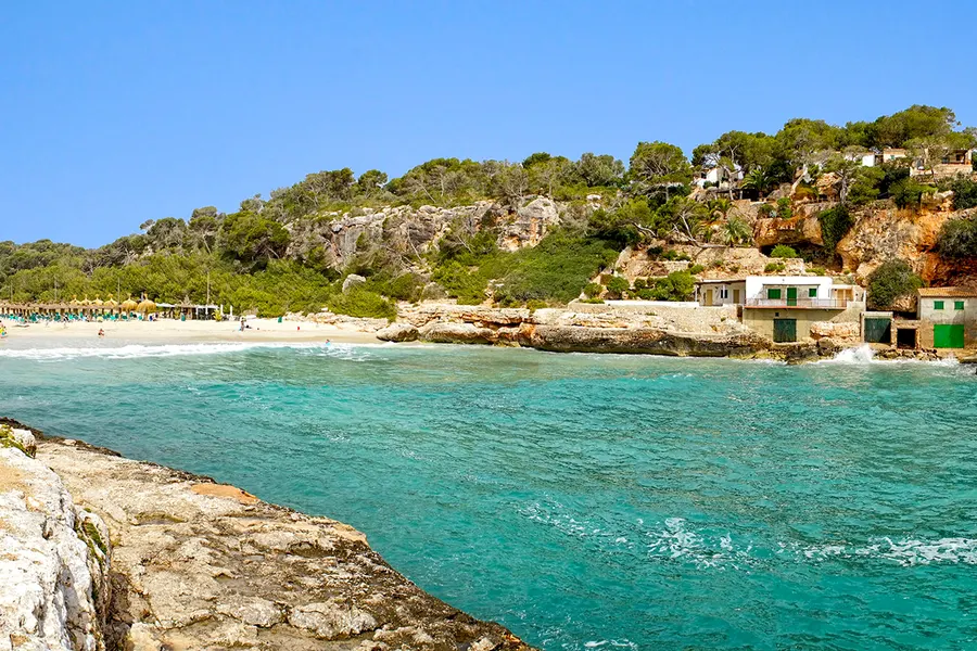 Blick vom Wasser her in die Cala Llombards mit türkisfarbenem Flachwasser, hellem Fels und den Bootshäusern am rechten Rand.
