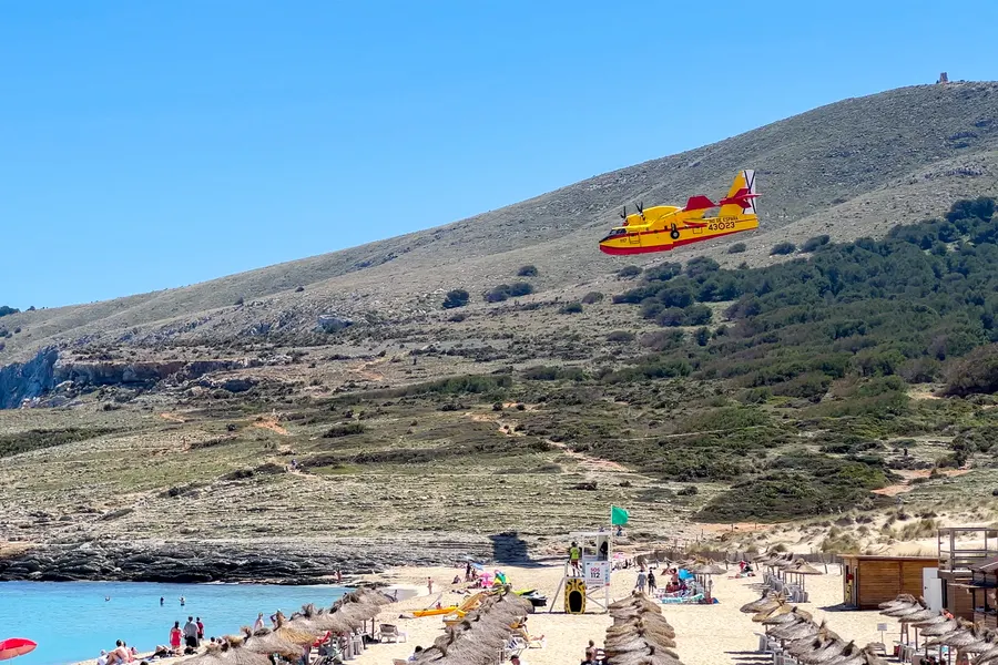 Gelb-rotes Löschflugzeug im Tiefflug über dem kahlen Hang hinter der Cala Mesquida, darunter der Strand mit Schirmen und Badegästen.
