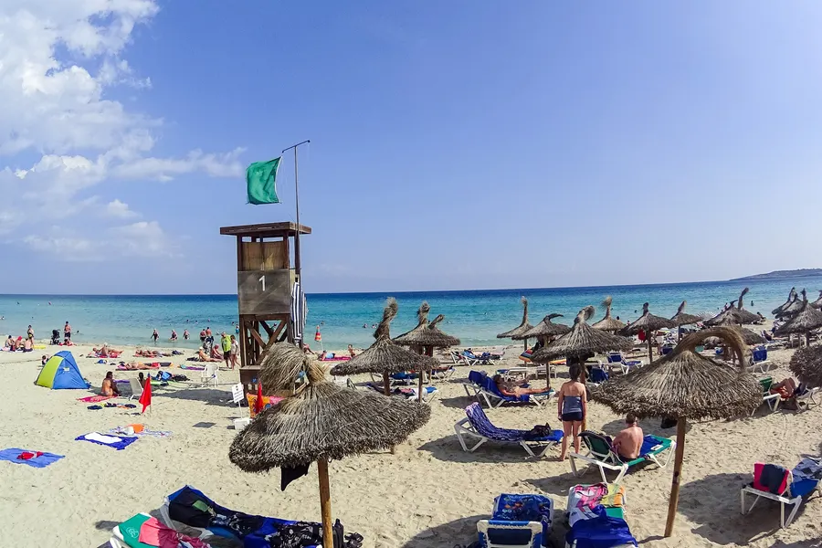 Sandstrand von Cala Millor mit Reihen von Strohschirmen, Liegen, Badegästen und einem hölzernen Rettungsturm.