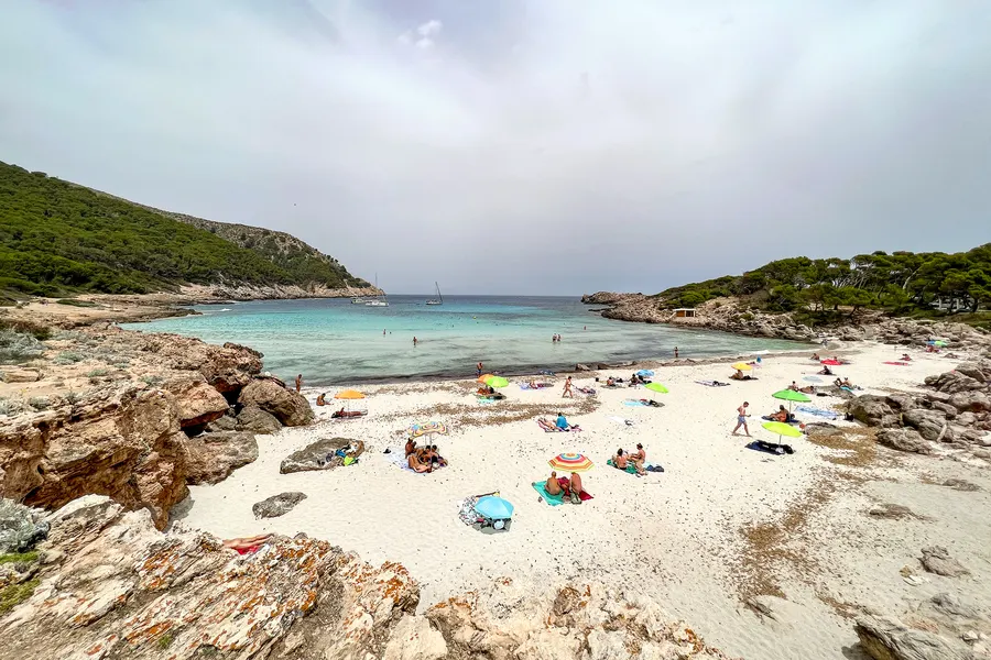 Blick von einem Felsvorsprung auf den schmalen Sandstrand der Cala Moltó mit einzelnen Sonnenschirmen und türkisfarbenem Wasser.