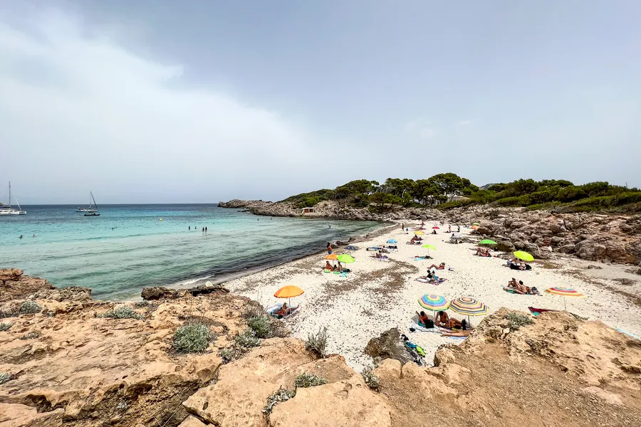 Einzelne Badegäste mit bunten Handtüchern auf dem Sand der Cala Moltó, davor sehr klares flaches Wasser über hellem Grund.