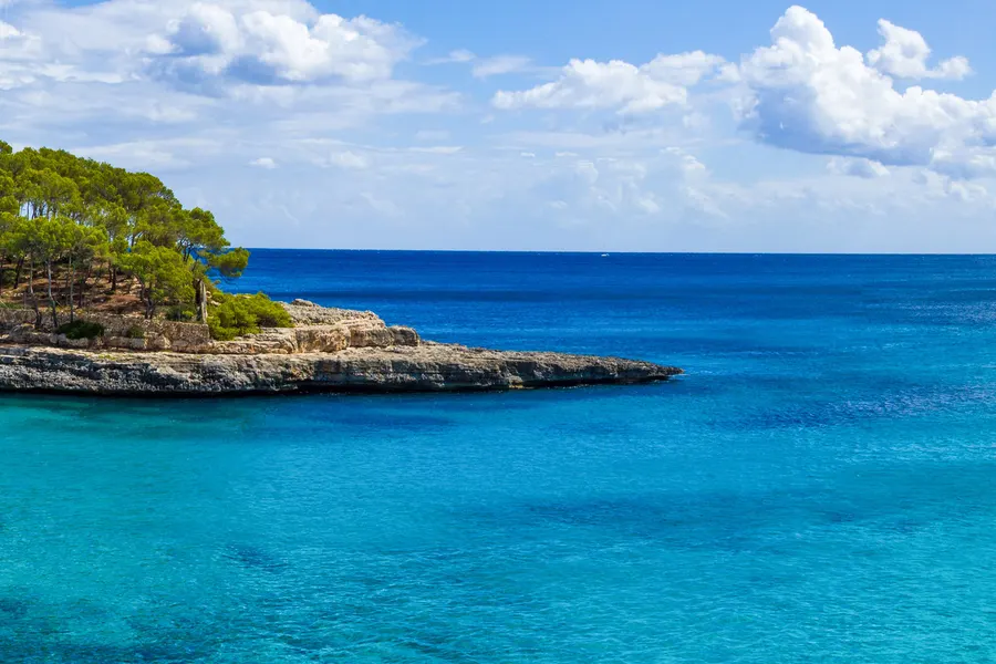 Flache helle Felsplatten am Ufer der Cala Mondragó mit sehr klarem türkisfarbenem Wasser und dahinter der offenen See.