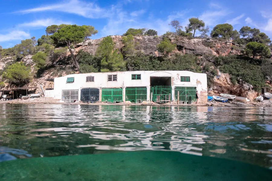 Weiß getünchtes Bootshaus mit grünen Toren direkt am Wasser der Bucht, davor klares flaches Meer und die Felswand dahinter.