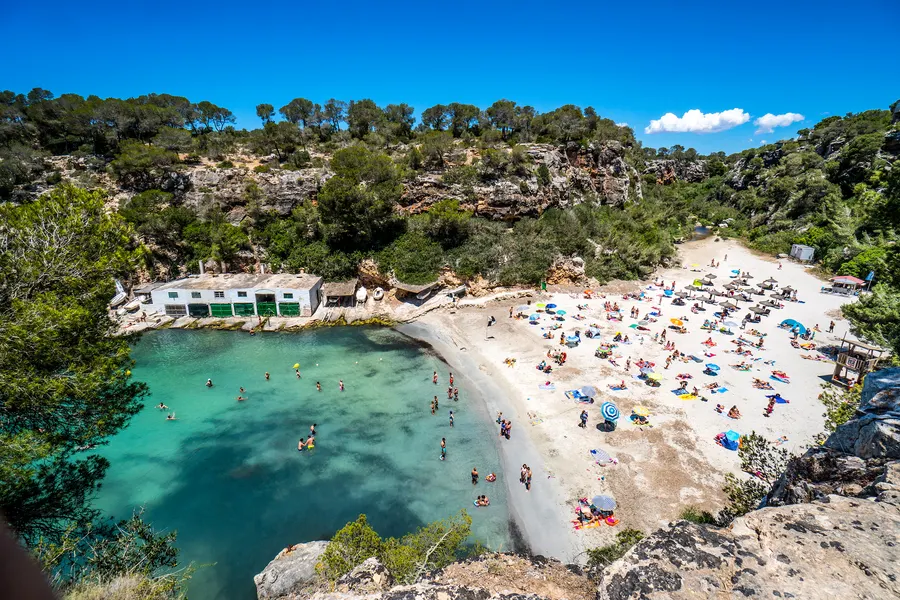 Der Sandstrand von Cala Pi im Sommer, dicht mit Handtüchern und Sonnenschirmen belegt, davor Badende im flachen türkisfarbenen Wasser.
