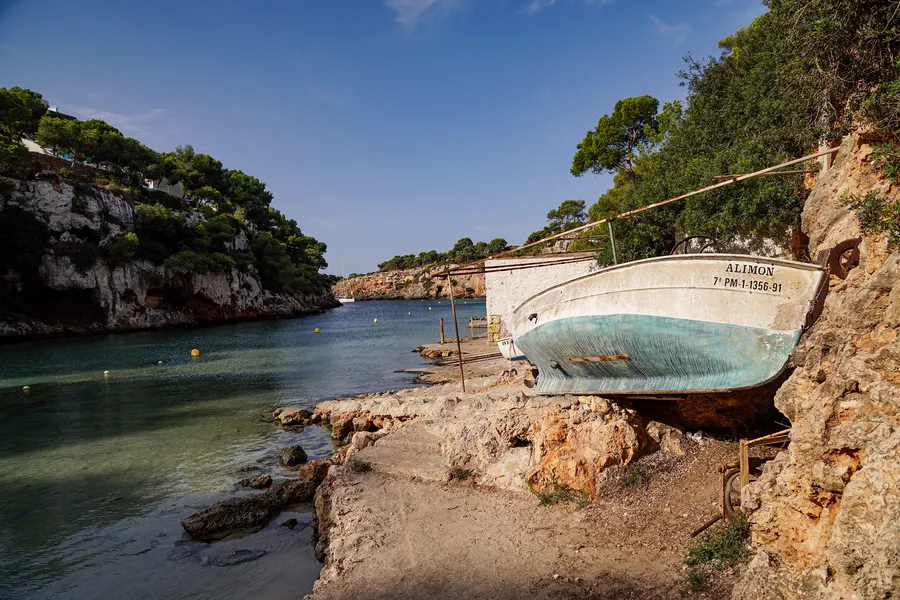 Ein weiß-türkis gestrichenes Holzboot mit dem Namen ALIMON steht auf einem Gestell am Felsufer von Cala Pi, davor die schmale Bucht mit Bojen, links die bewachsene Steilwand.
