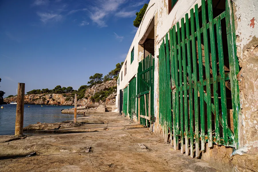Verwitterte Bootshäuser in Cala Pi mit grün gestrichenen Lattentoren und abgeplatztem Putz, davor die betonierte Slipbahn, links die Bucht mit einem Segelboot.

